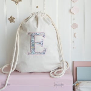 May include: A cream-coloured drawstring bag featuring a floral "E" appliqué. The bag has thick rope straps and is placed on a pink box. Gold glitter stars and pink cloud cutouts decorate the white wall.