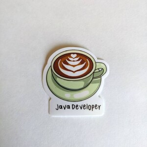 Może przedstawiać: Naklejka przedstawiająca filiżankę kawy z latte art w odcieniach brązu i zieleni na spodku. Pod spodem znajduje się napis "Java Developer". Naklejka jest na białym tle.