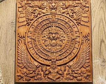 May include: A detailed, square, wooden carving of an Aztec calendar. The brown carving is on a light wood table, with a cup of tea, a white blanket, and a bundle of lavender. The carving has a central circular design with intricate details.