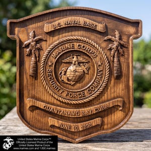May include: A wooden plaque with the U.S. Naval Base and Marine Corps emblems. The plaque features the text "Guantanamo Bay, Cuba" and "June 10, 1898." Two palm trees flank the central emblem. The plaque is a shield shape and has a dark wood finish.