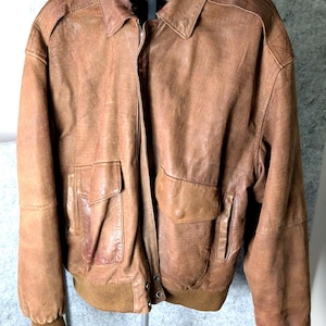 Könnte beinhalten: Braune Leder-Bomberjacke mit Kragen, zwei Vordertaschen und geripptem Saum und Bündchen. Die Jacke hat einen Reißverschluss und Schulterdetails. Das Leder hat ein abgenutztes Aussehen.
