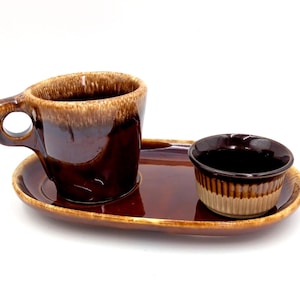 Pode incluir: Um conjunto de cerâmica composto por uma chávena esmaltada em castanho escuro com asa e uma pequena taça a condizer com bordos estriados, ambos sobre um tabuleiro oval. O conjunto apresenta um efeito de esmalte escorrido em dois tons.