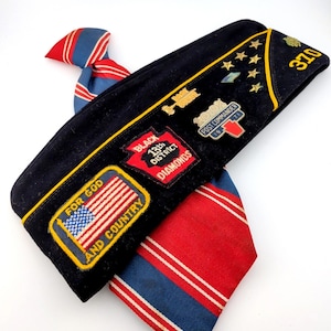 Op de afbeelding: Een zwarte militaire hoed met gouden rand, patches en spelden, waaronder een Amerikaanse vlag en de tekst "For God and Country". Een rood, wit en blauw gestreepte stropdas is eronder gedrapeerd. De hoed heeft het nummer "370".