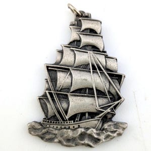Puede incluir: Colgante plateado con forma de velero. El colgante presenta representaciones detalladas de las velas, el aparejo y el casco del barco, con el barco navegando sobre agua ondulada. Un pequeño lazo en la parte superior para sujetarlo a una cadena.