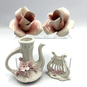 Może przedstawiać: Dwie ceramiczne rzeźby róż z jasnoróżowymi i białymi płatkami, mały biały ceramiczny dzbanek z różowym akcentem kwiatowym oraz biała ceramiczna figurka liry z różowymi detalami liści, wszystko o błyszczącym wykończeniu i gładkiej powierzchni.
