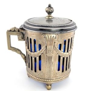 May include: An antique silver-toned cup with a lid and handle, featuring a blue interior. The cup has an ornate design with vertical bars and decorative elements. The lid is topped with a small finial.