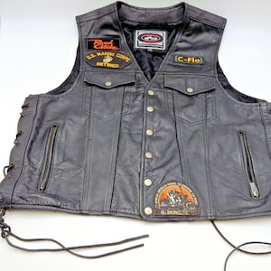 May include: A black leather motorcycle vest featuring a front button closure, two chest pockets, and side lace-up details. Patches include Road Glide, U.S. Marine Corps Retired, C-Flo, and an African American Riders Celebrating 130 Years Ironelite patch.