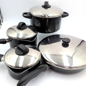 Könnte beinhalten: Ein Set aus vier schwarzen und silbernen Kochutensilien. Das Set umfasst einen Kochtopf, einen Topf mit zwei Griffen und eine Bratpfanne. Jedes Teil hat einen silbernen Deckel mit einem schwarzen Griff. Das Kochgeschirr steht auf weißem Hintergrund.