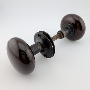Könnte beinhalten: Ein Vintage-Türknauf mit dunkelbraunem, glänzendem Finish. Der Knauf hat an jedem Ende eine ovale Form, die durch eine Metallstange und schwarze kreisförmige Platten verbunden ist. Die Hardware besteht wahrscheinlich aus Keramik und Metall.