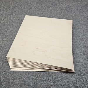 May include: A stack of light-colored wooden boards, possibly plywood, with visible wood grain. The boards are rectangular and appear to be new. The edges of the boards are visible, showing their thickness. The boards are stacked on a gray carpet.