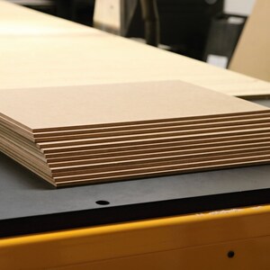 May include: A stack of rectangular, light brown wooden panels. The panels are neatly arranged on a black surface, with a larger wooden sheet in the background. The edges of the panels are visible, showing their thickness. The image suggests a woodworking or crafting project.