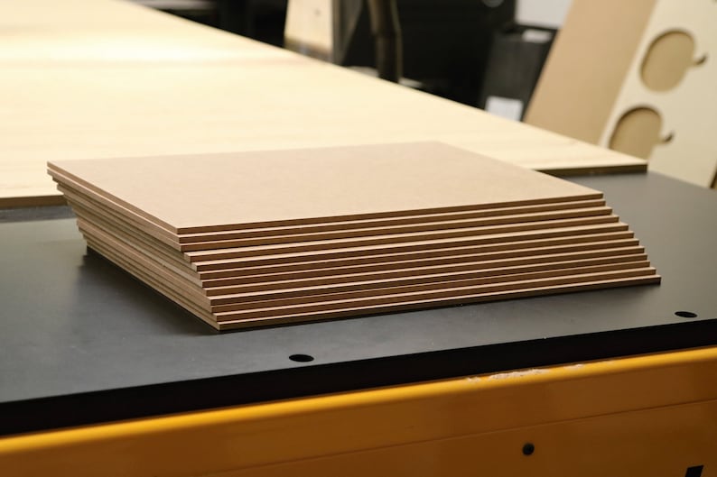 May include: A stack of rectangular, light brown wooden panels. The panels are neatly arranged on a black surface, with a larger wooden sheet in the background. The edges of the panels are visible, showing their thickness. The image suggests a woodworking or crafting project.