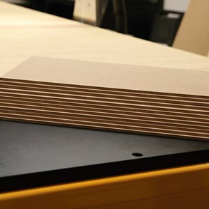 May include: A stack of rectangular, light brown wooden panels with dark brown edges. The panels are neatly arranged, revealing the layered construction. Additional wooden sheets are visible in the background, suggesting a woodworking project.