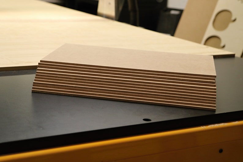 May include: A stack of rectangular, light brown wooden panels with dark brown edges. The panels are neatly arranged, revealing the layered construction. Additional wooden sheets are visible in the background, suggesting a woodworking project.