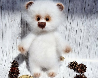 Puede incluir: Un oso de peluche blanco y esponjoso con detalles marrones en las orejas, la nariz y las patas. El oso tiene ojos oscuros y una pequeña etiqueta en el costado. El texto "tascha_toys" es visible en la esquina inferior derecha, junto a piñas.