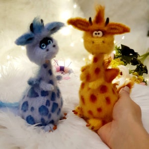 Puede incluir: Dos animales de peluche tejidos a crochet, un dragón azul y blanco y una jirafa amarilla y naranja, están sentados sobre una superficie blanca y peluda.
