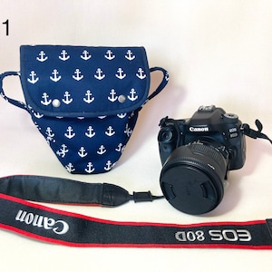 Op de afbeelding: Een zwarte Canon EOS 80D camera met een lens en een zwarte cameraband met rode bies en de tekst "Canon EOS 80D" erop gedrukt. De camera ligt naast een blauwe cameratas met een wit ankerpatroon.