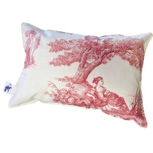 Può includere: Un cuscino bianco con un disegno floreale rosso che presenta un albero e una donna seduta sotto. Il cuscino ha un bordo bianco e un piccolo cartellino nero con un logo di cavallo bianco.