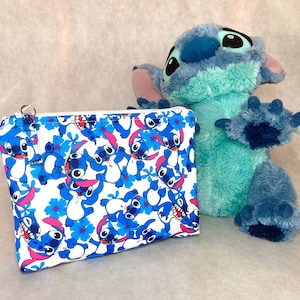 Può includere: Una pochette bianca con cerniera con un motivo blu e bianco di personaggi Stitch del film Disney Lilo & Stitch. La pochette è accanto a un giocattolo di peluche Stitch blu e verde.