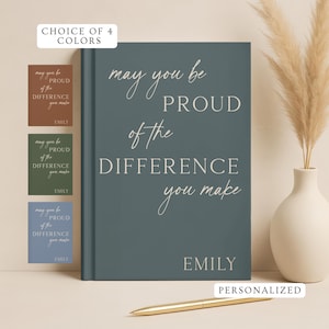 May include: A dark teal hardcover journal featuring the white text "may you be PROUD of the DIFFERENCE you make" and the name "EMILY" at the bottom. The image includes a gold pen and small color swatches showing four available color options.