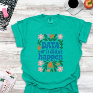 May include: Teal t-shirt featuring the phrase "DATA or it didn't happen" in blue, framed by a floral design with pink, orange, and green details. The t-shirt is a solid teal colour.