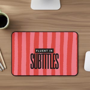 May include: A rectangular mousepad with a red and pink striped pattern. The mousepad features the text "FLUENT IN SUBTITLES" in black, bold letters. The mousepad is on a wooden desk, with a coffee mug and computer accessories visible.