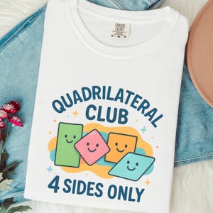 May include: White t-shirt with the text "QUADRILATERAL CLUB" and "4 SIDES ONLY" in blue. The design features smiling geometric shapes in green, pink, orange, and blue. The shirt is on a denim surface.