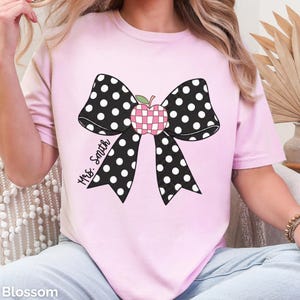 May include: A light pink t-shirt featuring a large black bow with white polka dots. In the centre of the bow is a pink and white checkered apple graphic. The text "Mrs. Smith" is printed in black cursive script on the left side of the bow.