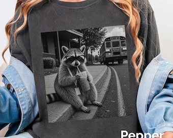 May include: A charcoal gray crewneck sweatshirt featuring a black and white graphic print of a raccoon wearing glasses, holding a drink with a straw, and sitting on a curb near a school bus. The word Pepper is printed in white at the bottom right.