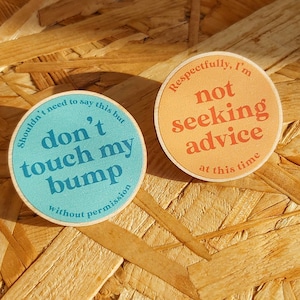 May include: Two round wooden signs with messages about pregnancy. The blue sign says "Shouldn't need to say this but don't touch my bump without permission". The orange sign says "Respectfully, I'm not seeking advice at this time".