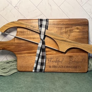 May include: A rectangular wooden cutting board with a handle and a curved wooden bread knife, both tied together with a black and white buffalo check ribbon. The board is engraved with the text Thankful Blessed & BREAD-OBSESSED.