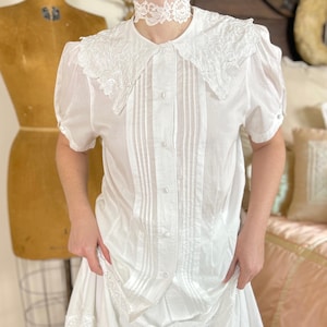 May include: A white short-sleeved blouse featuring a large embroidered collar, vertical pleats along the front button placket, and puff sleeves. The blouse is paired with a matching white skirt with lace trim and a white lace choker necklace.