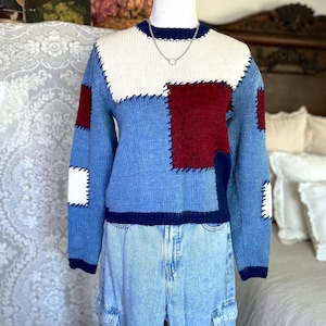 以下が含まれることがあります： 青、白、バーガンディの色合いのパッチワークセーター。セーターはクルーネックと長袖です。デザインは、目に見えるステッチが施された長方形のパッチを組み込んでいます。セーターはマネキンに展示され、ライトウォッシュデニムカーゴパンツと組み合わせています。