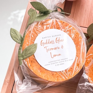 May include: A round bar of orange-colored soap wrapped in clear plastic with a white label that reads "EARTHY QUEENS Goddess Glow Turmeric & Lemon HANDCRAFTED ORGANIC LEMON SCENTED GOATS MILK SOAP 5 OZ (142g)"