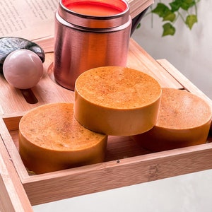 May include: Three round orange-colored soap bars stacked on top of each other on a wooden tray. A rose gold container with a red lid is in the background.