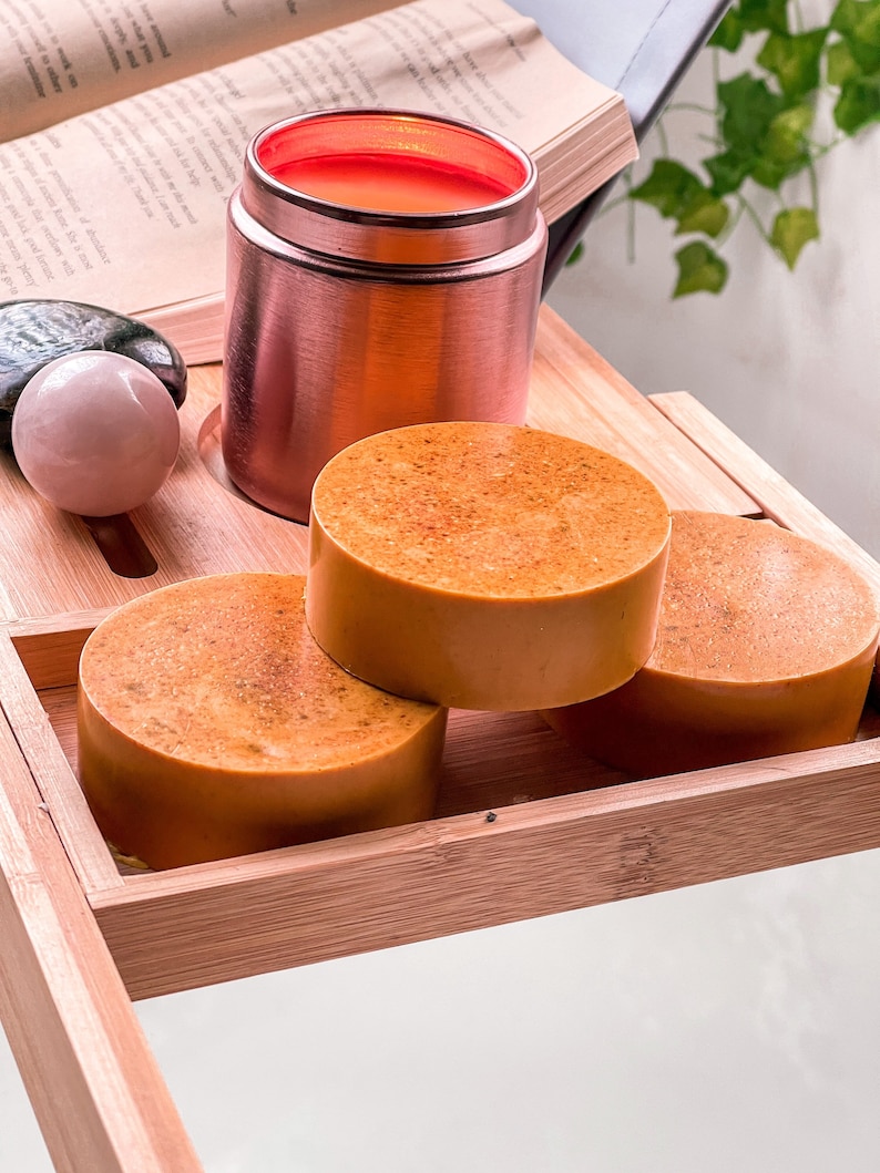 May include: Three round orange-colored soap bars stacked on top of each other on a wooden tray. A rose gold container with a red lid is in the background.