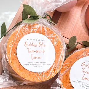 May include: Two round bars of handmade goat milk soap wrapped in clear plastic. The soap is a vibrant orange color and has a white label that reads "Goddess Glow Turmeric & Lemon" and "Handcrafted Organic Lemon Scented Goats Milk Soap 5 oz (142g)".