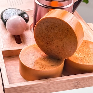 May include: Three round bars of orange-colored soap on a wooden tray. The soap is stacked on top of each other. The tray is made of bamboo and has a natural wood finish.
