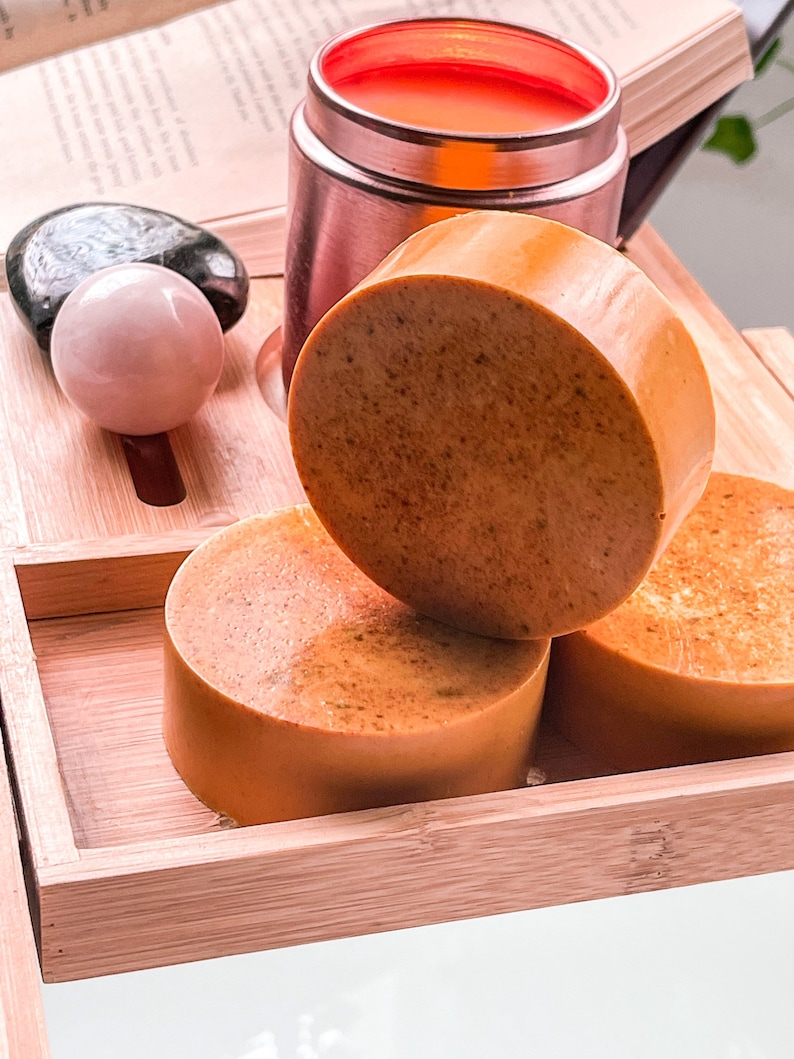 May include: Three round bars of orange-colored soap on a wooden tray. The soap is stacked on top of each other. The tray is made of bamboo and has a natural wood finish.