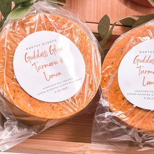 May include: Two round bars of handmade organic goats milk soap wrapped in clear plastic with a white label that reads "Earthy Queens Goddess Glow Turmeric & Lemon Handcrafted Organic Lemon Scented Goats Milk Soap 5 oz (142g)"