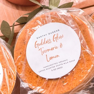 May include: A round, orange-colored goat milk soap bar with a white label that reads "Goddess Glow Turmeric & Lemon" and "Handcrafted Organic Lemon Scented Goats Milk Soap 5 oz (142g)".