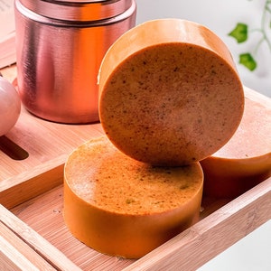 May include: Three round orange-brown soap bars on a wooden tray. The soap bars are stacked on top of each other.