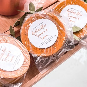 May include: Three bars of handmade soap wrapped in plastic. The soap is a bright orange color and has a label that says "Goddess Glow Turmeric & Lemon" with the words "Handcrafted Organic Lemon Scented Goats Milk Soap 5 oz (142g)".