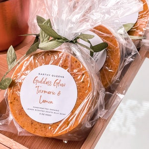 May include: Two round bars of orange-colored goat milk soap wrapped in clear plastic with a green leaf tied around them. The soap is labeled "Goddess Glow Turmeric & Lemon" and "Handcrafted Organic Lemon Scented Goats Milk Soap 5 oz (142g)"