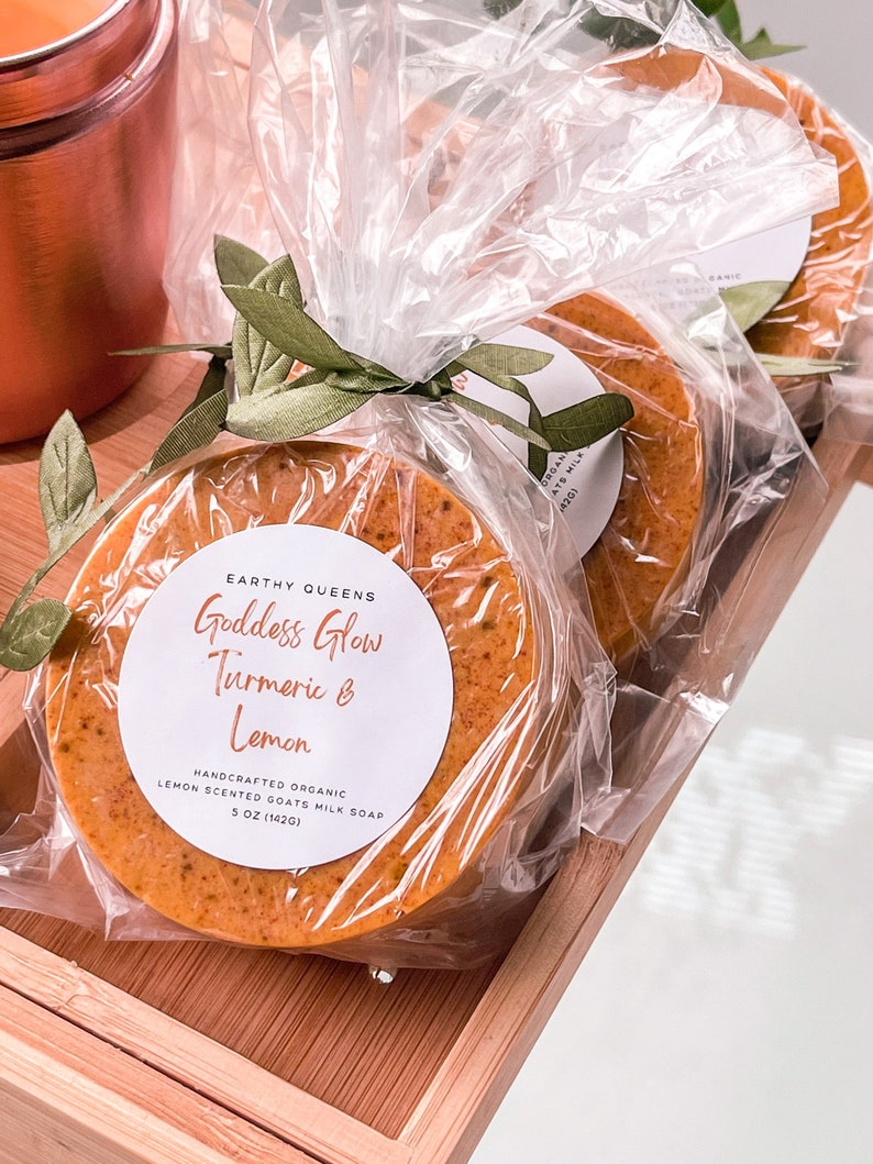 May include: Two round bars of orange-colored goat milk soap wrapped in clear plastic with a green leaf tied around them. The soap is labeled "Goddess Glow Turmeric & Lemon" and "Handcrafted Organic Lemon Scented Goats Milk Soap 5 oz (142g)"
