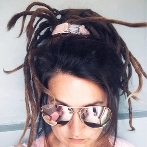 Könnte beinhalten: Eine Frau mit langen braunen Dreadlocks trägt eine roségoldene Aviator-Sonnenbrille. Ihre Haare sind mit einem rosa-silbernen Haargummi zurückgebunden.