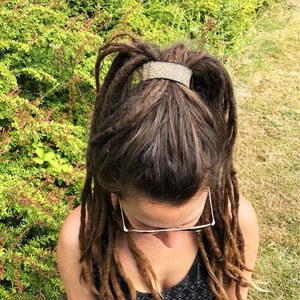 Könnte beinhalten: Eine Frau mit braunen Dreadlocks trägt ein schwarz-weiß gemustertes Kleid und eine Brille. Ihre Haare sind mit einem glitzernden silbernen Haargummi zurückgebunden.