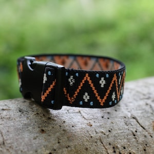 K&ouml;nnte beinhalten: Ein schwarzes Gummiband mit einem geometrischen Muster in Orange, Wei&szlig; und Blau. Das Band hat eine schwarze Kunststoffschnalle.