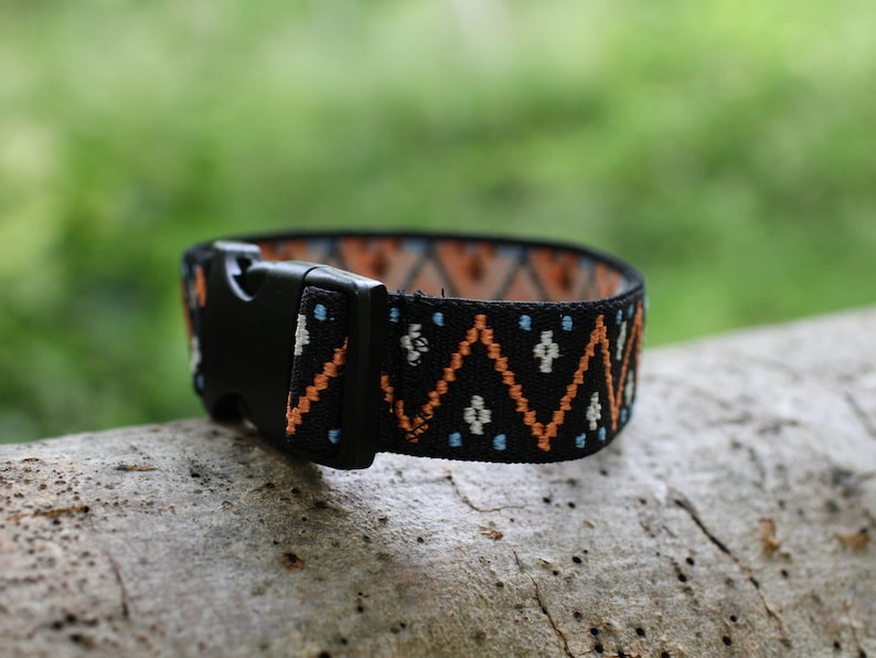 K&ouml;nnte beinhalten: Ein schwarzes Gummiband mit einem geometrischen Muster in Orange, Wei&szlig; und Blau. Das Band hat eine schwarze Kunststoffschnalle.