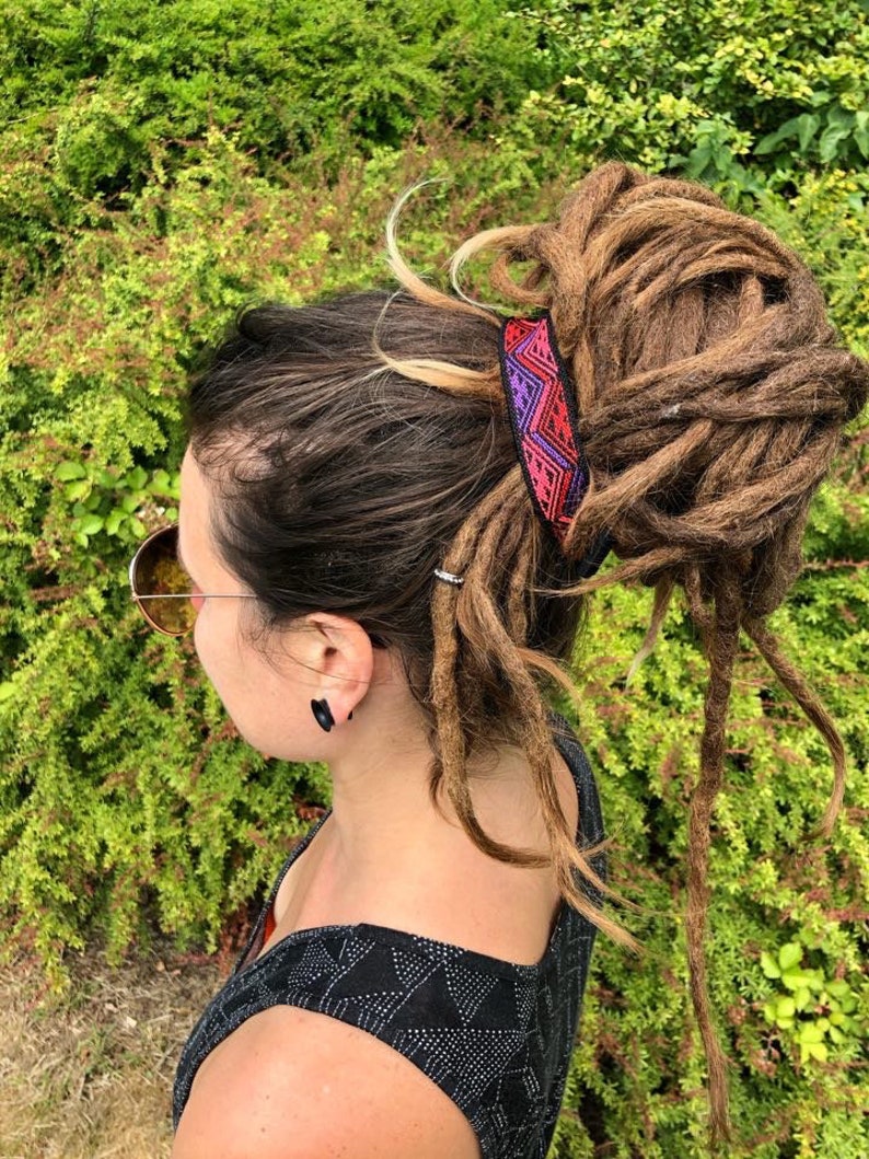 K&ouml;nnte beinhalten: Eine Person mit langen braunen Dreadlocks, die zu einem Dutt gebunden sind und mit einem bunten, gemusterten Haargummi befestigt sind. Das Haargummi hat ein geometrisches Muster in Pink, Lila und Schwarz.